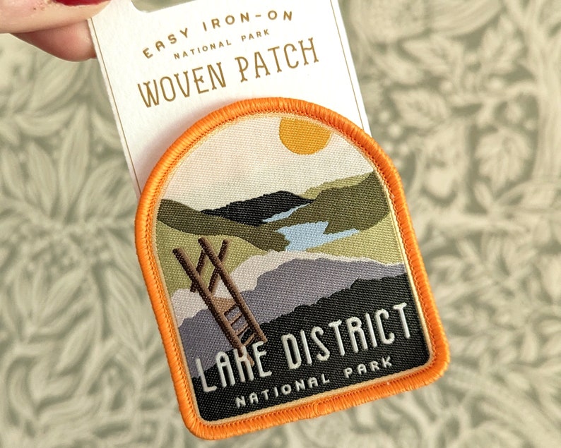 K&ouml;nnte beinhalten: Ein orangefarbener B&uuml;gel-Patch mit einer Landschaftszene eines Sees, Bergen und einer Leiter. Der Text "Lake District National Park" befindet sich auf dem Patch.