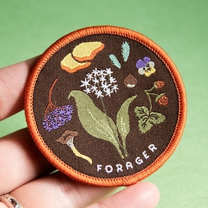 May include: Circular embroidered patch with a brown backdrop and orange edging. The patch displays embroidered plants and the word "FORAGER". The plants include mushrooms, leaves, and flowers.