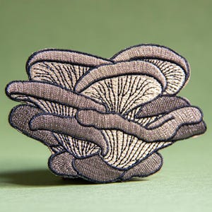 Peut inclure: Écusson brodé représentant un groupe de pleurotes. Le patch présente un design détaillé avec des nuances de gris et de beige, et des contours noirs. L'écusson champignon est sur un fond vert.