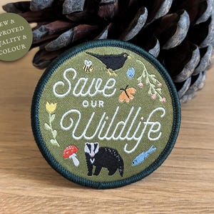 Puede incluir: Parche bordado verde con el texto "Save Our Wildlife" en letras cursivas blancas. El parche presenta un tejón, un mirlo, una mariposa, abejas y flores.