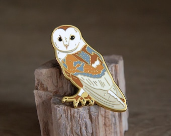 May include: A gold-colored metal pin in the shape of a barn owl. The owl has a white face, tan and brown wings with blue accents, and a gold outline. The pin is resting on a piece of wood.