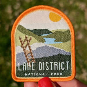 K&ouml;nnte beinhalten: Orangefarbener und wei&szlig;er gestickter Aufn&auml;her mit einer Landschaftszene eines Sees, Bergen und einer Leiter. Der Text "LAKE DISTRICT NATIONAL PARK" ist in Schwarz auf dem Aufn&auml;her aufgedruckt.