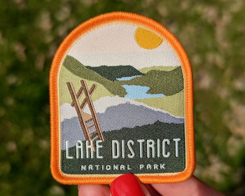 K&ouml;nnte beinhalten: Orangefarbener und wei&szlig;er gestickter Aufn&auml;her mit einer Landschaftszene eines Sees, Bergen und einer Leiter. Der Text "LAKE DISTRICT NATIONAL PARK" ist in Schwarz auf dem Aufn&auml;her aufgedruckt.
