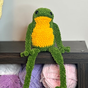 May include: A green crocheted frog with a yellow belly sitting on a shelf with balls of yarn in various colors.
