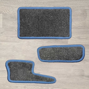 May include: A set of three dark grey floor mats with a textured surface and bright blue stitched edges. The set includes one large rectangular mat, one smaller rectangular mat with rounded corners, and one L-shaped mat with rounded edges.