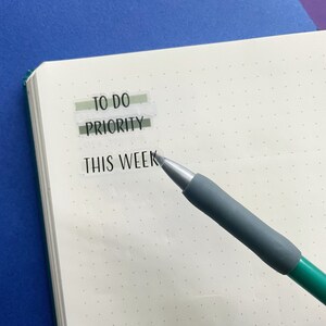 Op de afbeelding: Een wit notitieboek met een grijze pen erop. Het notitieboek heeft een gestippeld rasterpatroon en heeft drie transparante stickers op de pagina. De stickers zeggen "To Do", "Priority" en "This Week".