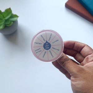 May include: A circular reading spinner in pink and white, featuring the text "My Next Read" at its centre. The spinner is segmented, with labels including "Mystery", "Fantasy", "Science Fiction", "Horror", "Romance", "Thriller", and "Fiction".