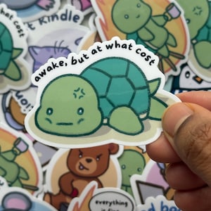 May include: A sticker featuring a cartoon turtle with a teal shell and a grumpy expression. The text "awake, but at what cost" arches above the turtle. Other stickers with cartoon animals and text are visible in the background.