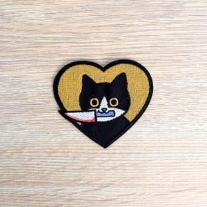 May include: A black and white cat with a red knife in its mouth, inside a yellow heart-shaped outline.