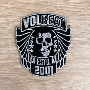 Op de afbeelding: Een zwart-wit geborduurde patch met het woord "VOLBEAT" boven een schedelontwerp. De patch bevat de tekst "ESTD. 2001" en vleugelachtige details. De patch ligt op een lichtbruin houten oppervlak.