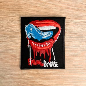 May include: Black rectangular patch with a stylized mouth design in red, white, and blue. The open mouth has red drips, and the text "FALLING IN REVERSE" is embroidered in white below. A music-themed accessory.