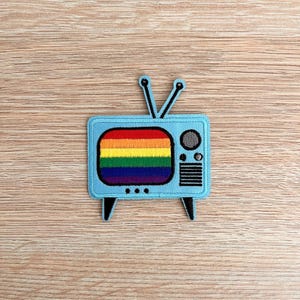 Puede incluir: Un parche bordado azul claro con forma de televisor retro. La pantalla muestra un diseño de arcoíris. El parche tiene antenas, patas y detalles negros.