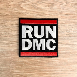 Op de afbeelding: Vierkante zwarte patch met de tekst "RUN DMC" in witte blokletters. De boven- en onderkant van de patch hebben een rode rand. De patch ligt op een lichtbruin houten oppervlak.