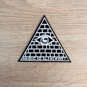 Puede incluir: Un parche bordado en blanco y negro que presenta un ojo que todo lo ve dentro de una pirámide con el texto "MDCCLXXVI" en la parte inferior.