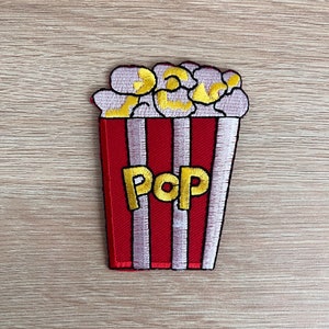 Puede incluir: Un cubo de palomitas de maíz a rayas rojas y blancas con la palabra "Pop" bordada en amarillo. El cubo está lleno de granos de palomitas de maíz.
