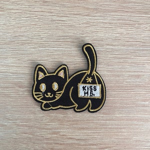 Könnte beinhalten: Schwarzer gestickter Bügelaufkleber mit einem Kätzchen, das ein Schild mit der Aufschrift "Kiss Me" auf dem Rücken trägt.