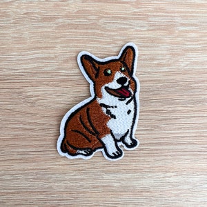 May include: Embroidered patch of a brown and white corgi dog with a happy expression, sitting with its tongue sticking out.