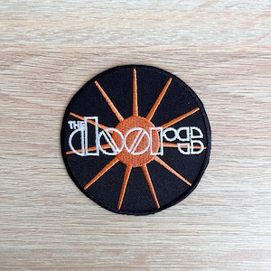Può includere: Una toppa ricamata nera e arancione con le parole "The Doors" in lettere bianche. La toppa presenta un design a raggiera stilizzato.