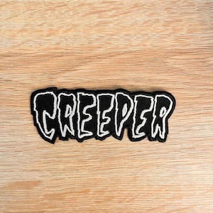 May include: A black embroidered patch featuring the word CREEPER in a jagged, white, stylized font. The lettering has a thick, uneven outline and a textured appearance, with the entire design bordered by a solid black stitched edge.