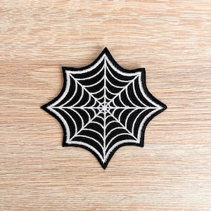 Op de afbeelding: Zwarte en witte geborduurde spinnenweb patch. Het web is wit en de achtergrond is zwart.