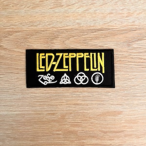 May include: A black rectangular patch featuring the words "LED-ZEPPELIN" in yellow, with four white symbols below. This patch, likely for clothing or bags, celebrates the iconic rock band Led Zeppelin.