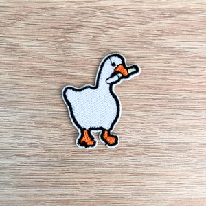 May include: White embroidered goose holding a carrot in its beak. The goose is standing on a light brown wooden surface.