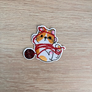 Peut inclure: Écusson brodé représentant un chat orange et blanc enroulé dans du fil rouge, avec une petite pelote de laine. Le chat a des oreilles et des pattes roses. L'écusson est posé sur une surface en bois brun clair.