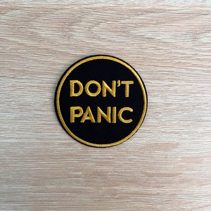 Könnte beinhalten: Ein schwarzer runder Patch mit goldener Umrandung und dem Text "DON'T PANIC" in goldenen Buchstaben.