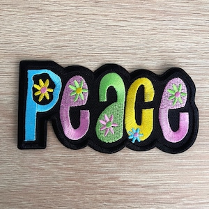 May include: Embroidered patch featuring the word "peace" in colourful bubble letters. The letters are outlined in black and decorated with flower designs. The patch has a black scalloped border.