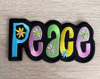 May include: Embroidered patch with the word "peace" in colorful bubble letters. The letters are outlined in black and decorated with flower designs. The patch has a black scalloped border.
