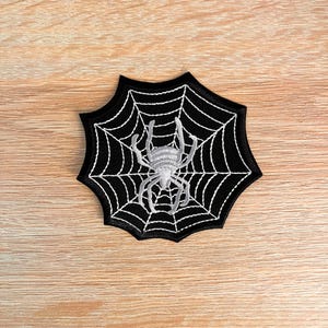 Puede incluir: Un parche bordado negro con forma de telaraña y una araña blanca cosida en el centro. El diseño de la red presenta líneas blancas radiantes que se conectan a un borde circular, creando un elemento gráfico detallado para aplicar en telas.