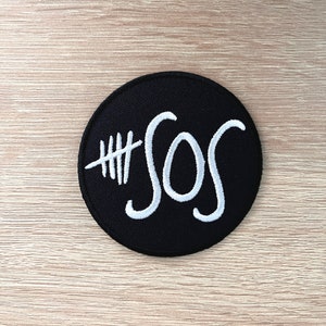 Peut inclure: Un patch rond noir avec une broderie blanche. Le patch présente le texte "#SOS" dans une police stylisée.