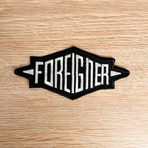 May include: A black and white embroidered patch featuring the word "FOREIGNER". The text is in capital letters and is set within a black diamond shape with white arrow details. The patch is designed with a bold, blocky font.