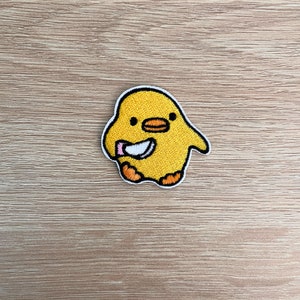 Op de afbeelding: Een geborduurde patch met een cartoonkuiken dat een mes vasthoudt. Het kuiken is geel met zwarte ogen en een zwarte snavel.