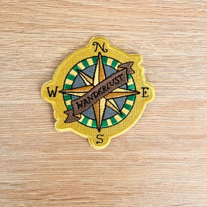 Op de afbeelding: Geborduurde patch met een kompasontwerp en het woord "WANDERLUST" op een banner. De patch heeft een gele rand, een groen en blauw kompasroos en gouden richtingspijlen. De letters N, W, E en S markeren de windrichtingen.