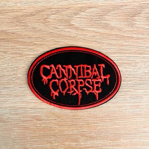 May include: An oval-shaped embroidered patch featuring a black background with a red border. The text CANNIBAL CORPSE is stitched in a stylized red font with a dripping effect across the center of the patch, creating a bold and high-contrast design.