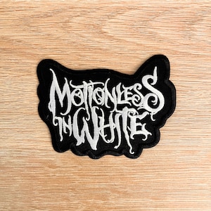 May include: A black embroidered patch featuring the words "Motionless in White" in white, stylized lettering. The patch has an irregular outline and is set against a light wood grain surface. The text is in a gothic font.