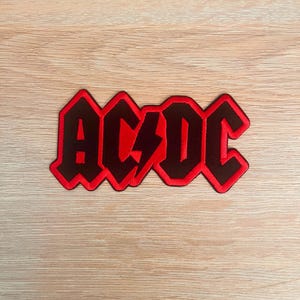 Pode incluir: Patch bordado preto e vermelho com o texto "AC/DC" e um raio. O patch tem uma borda vermelha. O fundo é de grão de madeira clara.