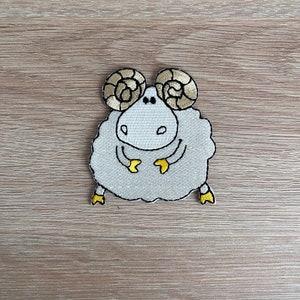 Op de afbeelding: Een witte geborduurde patch van een cartoon ram met gele hoorns en hoeven. De ram staat met zijn benen licht uit elkaar en zijn hoofd lichtjes gekanteld.