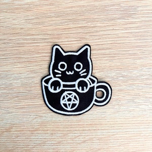 Könnte beinhalten: Schwarzer gestickter Bügel-Patch mit einem schwarzen Kater, der in einer Kaffeetasse mit einem Pentagramm darin sitzt.