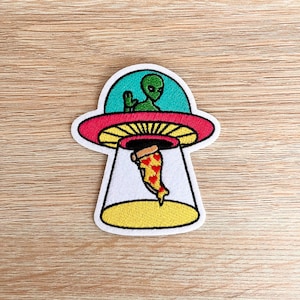 May include: An embroidered patch depicting a green alien in a flying saucer, holding a slice of pizza. The saucer is red, yellow, and blue, and the pizza is red, yellow, and orange.