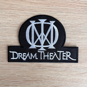 以下が含まれることがあります： ドリーム・シアターのロゴが刺繍された黒と白のパッチ。ロゴは円の中にスタイリッシュな「M」が描かれています。「Dream Theater」という文字がロゴの下にあります。