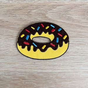 May include: A yellow donut-shaped patch with a chocolate frosting design and colorful sprinkles. The patch has a yellow border and is designed to be sewn or ironed onto fabric. This patch is perfect for adding a touch of fun to clothing or accessories.