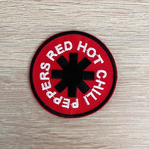 May include: A circular, embroidered patch with the text "PEPPERS RED HOT CHILI" in white, arched around a black asterisk-like symbol. The patch has a red background and a black border.