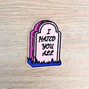 Puede incluir: Parche bordado rosa y morado con forma de lápida. El parche presenta el texto "I HATED YOU ALL" en escritura negra. Hierba azul en la parte inferior de la lápida.