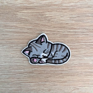 May include: An embroidered patch of a sleeping cat, with grey and white fur and pink ears and paws.