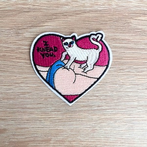 May include: Embroidered patch in a heart shape with a white border. The patch features a cartoon cat with blue eyes and the text "I KNEAD YOU." The cat is on a pink background.