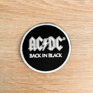 Puede incluir: Un parche redondo negro con borde blanco. El parche presenta el nombre de la banda "AC/DC" en blanco, letras estilizadas con un rayo y el texto "BACK IN BLACK" debajo.