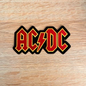 May include: An embroidered patch featuring the text AC/DC in a bold, red font with a yellow border. The letters are outlined in black, and a red lightning bolt symbol separates the AC and DC, all set against a black background shape.