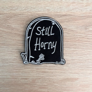 Puede incluir: Un parche negro en forma de lápida con texto bordado en blanco que dice "Still Horny". El parche tiene un borde blanco y un pequeño gráfico de flores en la parte inferior.